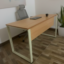 Modern wooden office desk with a light oak finish top, white metal U-shaped legs, and a matching wooden modesty panel, viewed from a high-angle perspective in a bright office setting.