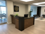 Modern L-shaped wooden reception desk with walnut finish and black privacy panel, professional office front desk furniture on light wood flooring.