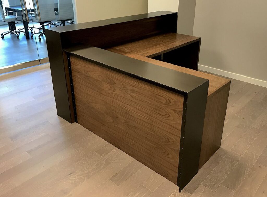 Modern L-shaped wooden reception desk with walnut finish and black privacy panel, professional office front desk furniture on light wood flooring.