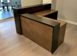 Modern L-shaped wooden reception desk with walnut finish and black privacy panel, professional office front desk furniture on light wood flooring.