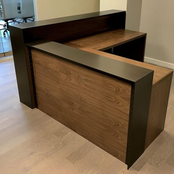 Modern L-shaped wooden reception desk with walnut finish and black privacy panel, professional office front desk furniture on light wood flooring.