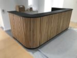 Modern reception desk featuring vertical light wood slats, curved edges, and a matte black work surface for a luxury office lobby.