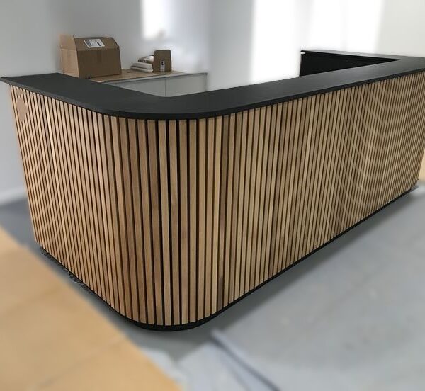 Modern reception desk featuring vertical light wood slats, curved edges, and a matte black work surface for a luxury office lobby.